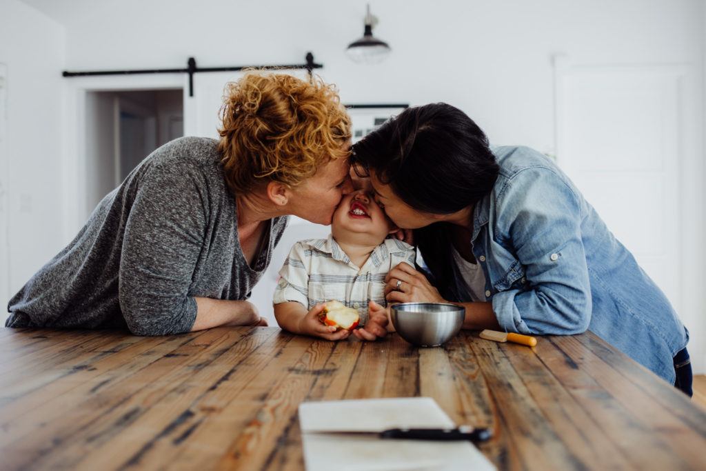 Two moms kiss their toddler son in their kitchen in Los Angeles, CA