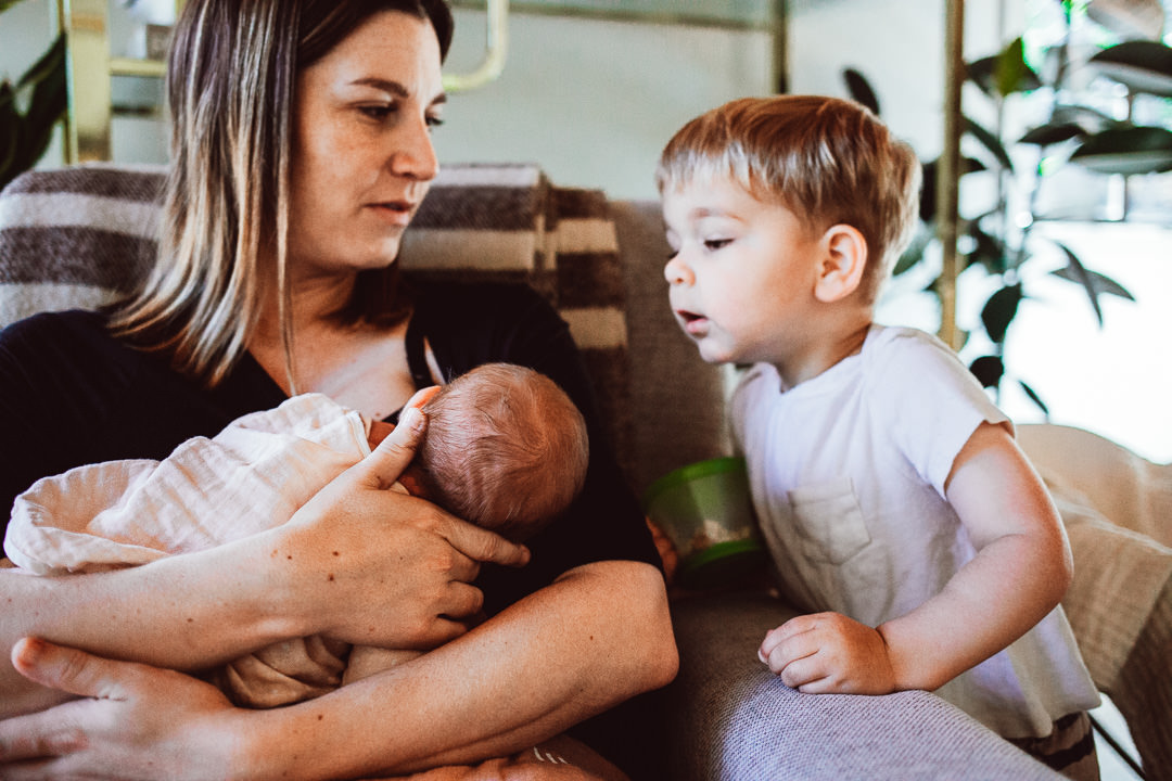 Mom Holding Newborn Baby With Toddler Son Looking Over Shoulder