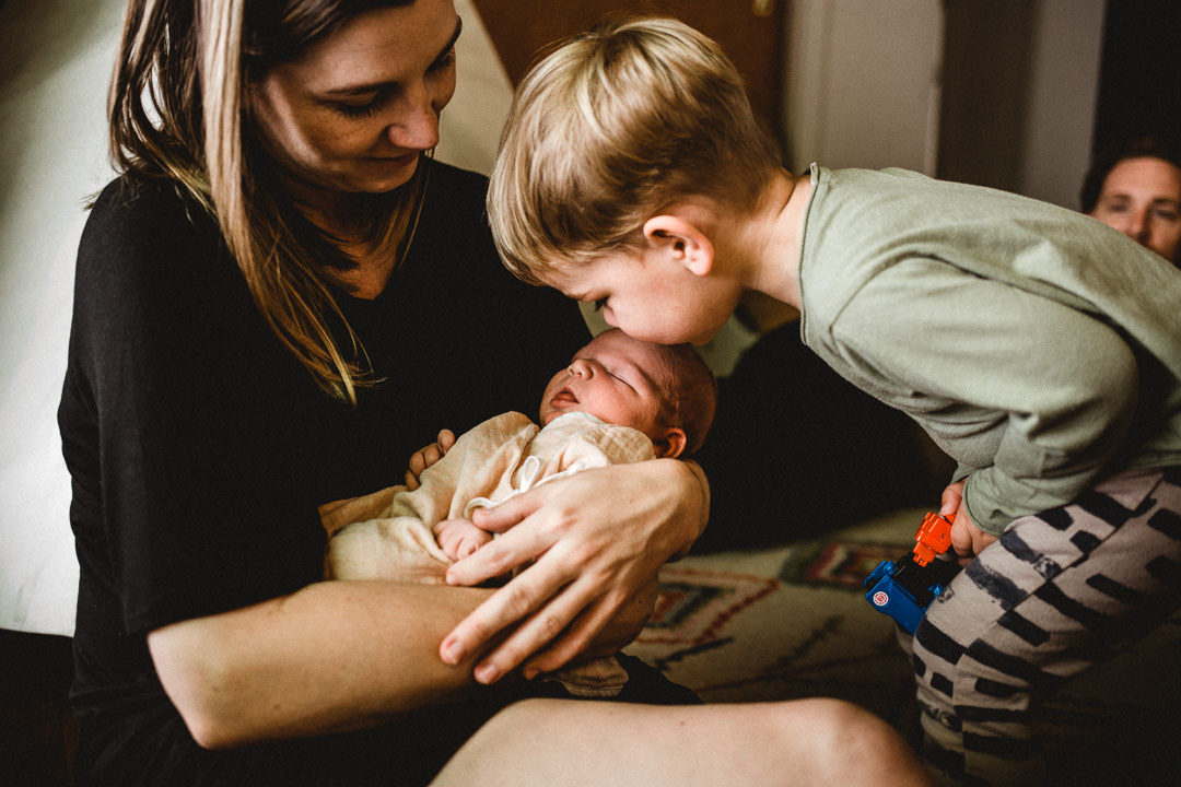 Toddler Kissing Newborn Baby's Head While Mom Holds Baby