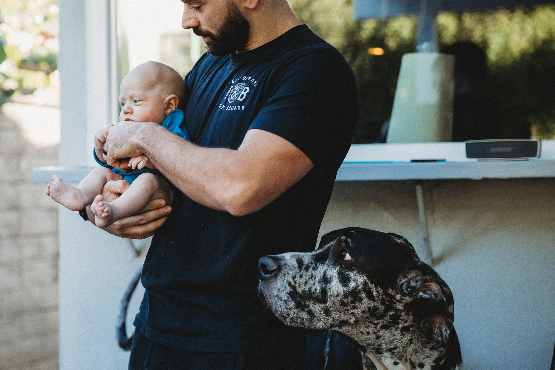 Dad holds baby while Great Dane looks on, outside their home in Los Angeles, CA