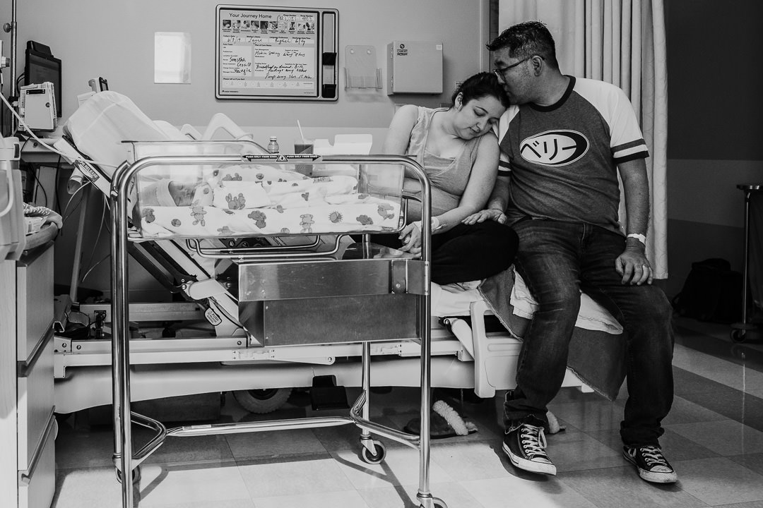 Dad Kissing Mom While Newborn in Bassinet in Hospital Black and White Photo