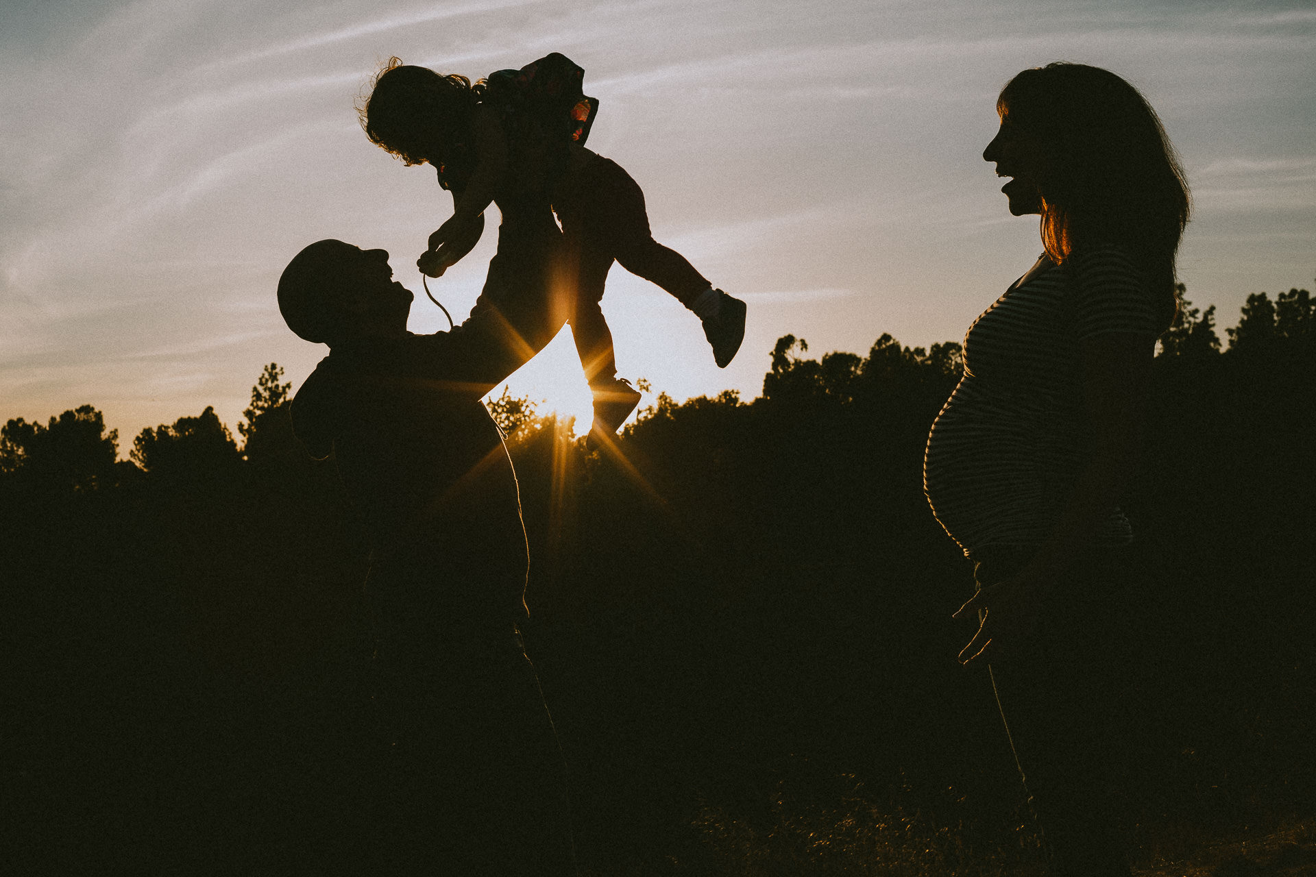 Dad Holding Toddler Up in Air At Dusk With Pregnant Mom Looking On