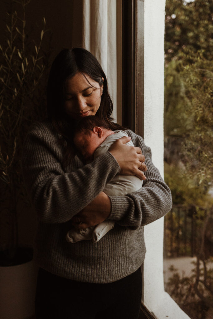 Mom Snuggles Newborn Baby By Window in House