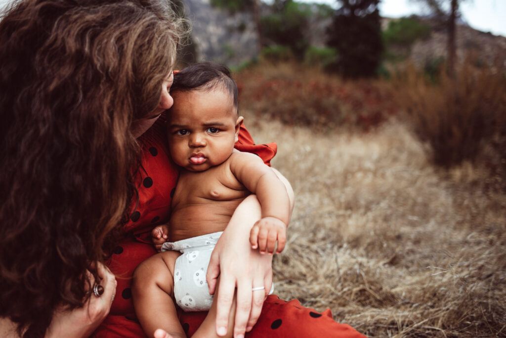 Baby In Diaper Looking At Camera While Being Held By Mom in Red Dress