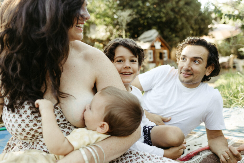 Baby nursing in mom's arms while whole family sits together outside in Pasadena, CA