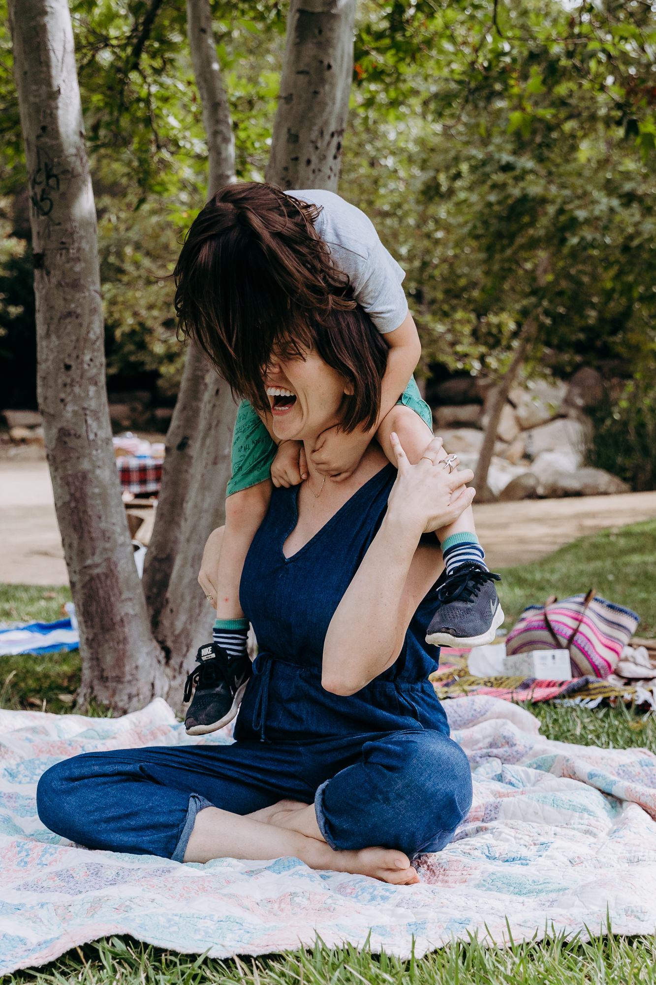 Young Kid Climbs On Top of Moms Shoulders While Sitting Outside On Picnic Blanket